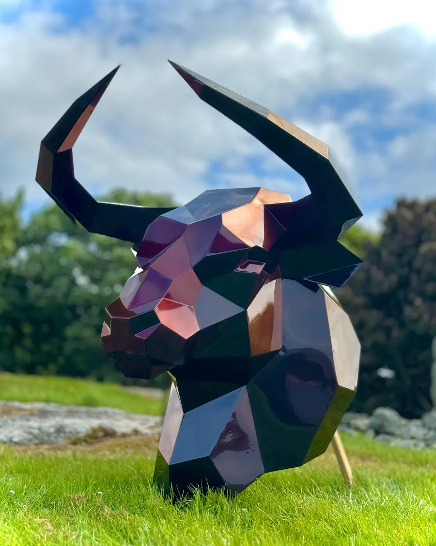 Low-poly steel bull head sculpture with an iridescent finish standing on grass